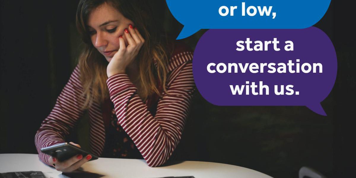 img94joktmu78493 Woman using her phone in a dark room. Text bubbles say: If you're feeling anxious or low, start a conversation with us. White text on a blue banner reads: Text HEAR to 85258 Free confidential support 24/7. NHS Peterborough and Cambridgeshire, and in partnership with shout logos are on the bottom right corner.